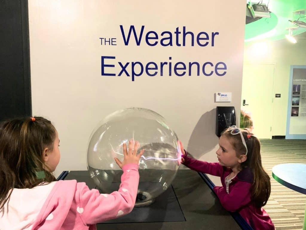 Two girls are standing in a room next to a phrase on the wall titled "the Weather Experience", with palms on a clear plasma globe that is sending out electricity to their hands.