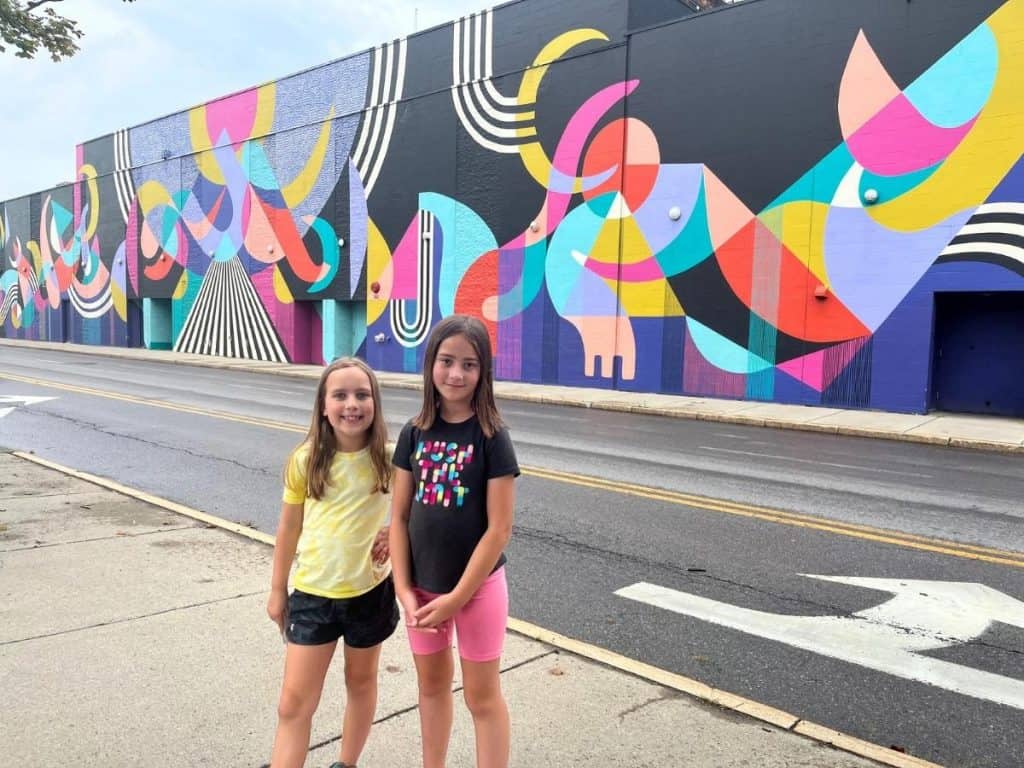 Kids can see all types of murals or interesting attractions on family trips, which can be easier to plan than you think! Two girls in t-shirts and shorts stand across the street from a building with a colorful mural of shapes and swirls in pink, yellow, green, purple, black, and white.