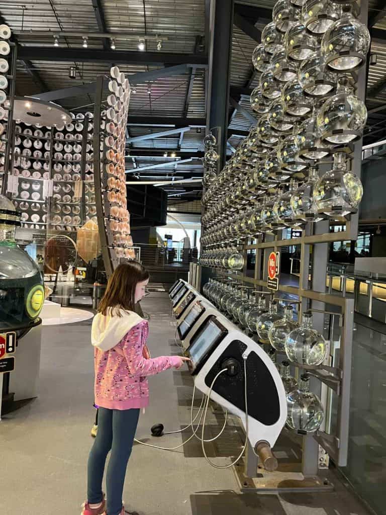 A girl in a pink sweatshirt stands in front of a series of free-standing computer terminals below hundreds of circular glass water jugs on the wall.