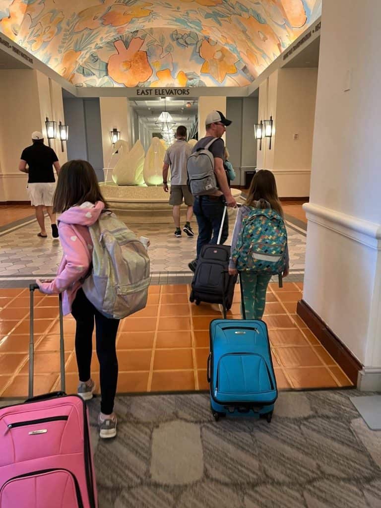 Three family members carry minimal luggage during a trip along with minimal stuffed animals. Older kids carry their own suitcases. Viewing 2 children and a man walking away inside a hotel with a fountain. Each carries a backpack and rolling luggage.
