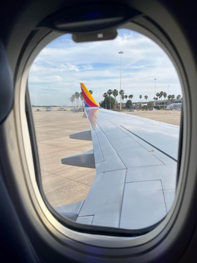 An airplane window view shows an airplane wing with blue, red, and yellow Southwest colors, the runway, and palm trees in the background.
