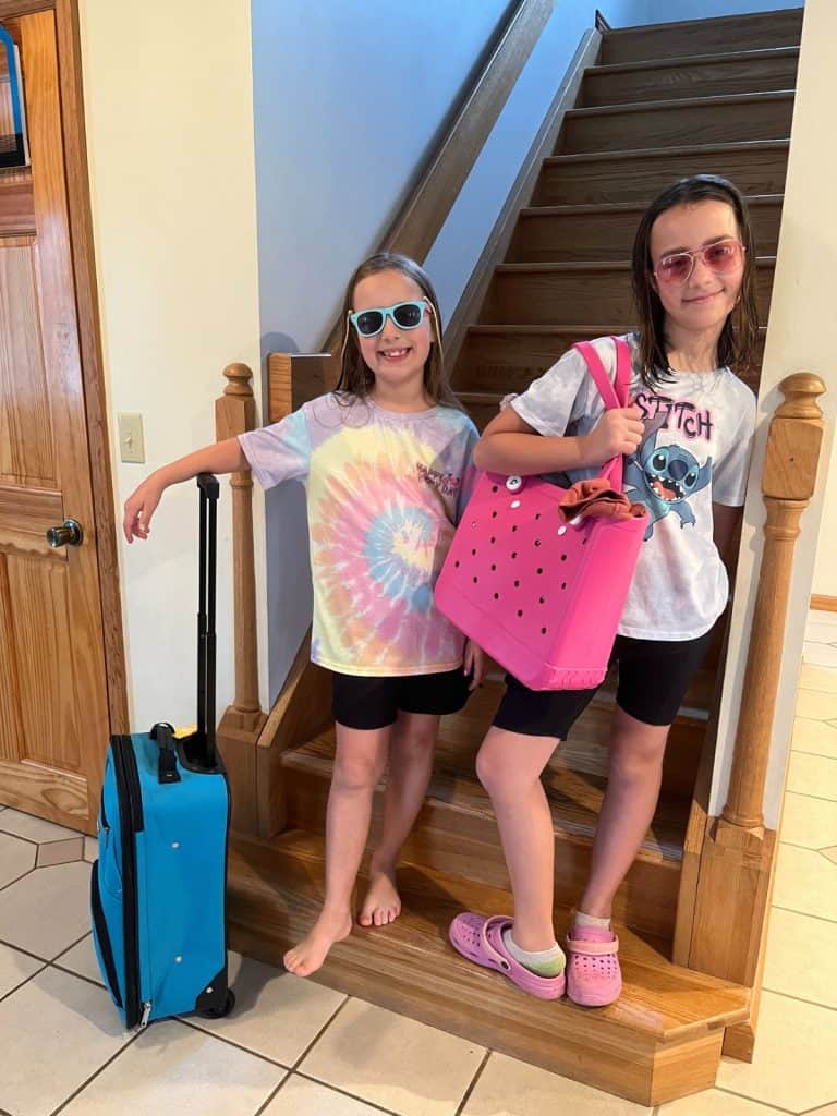 Two girls stand on the stairs with luggage, getting excited before a family vacation they're going to take. Two girls with sunglasses, colorful t-shirts, and shorts, stand on a set of stairs with a rolling suitcase and summer pink shoulder bag.