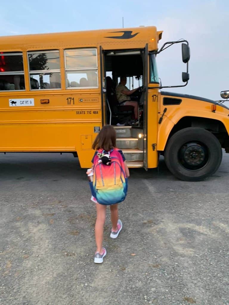 A girl with a rainbow bookbag walks toward a yellow school bus.