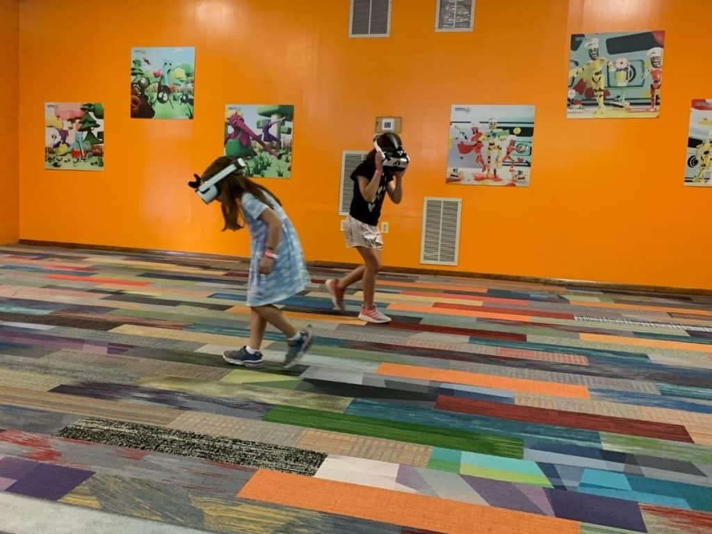 Two girls have Virtual Reality headsets on at an orange theme park room with kids' pictures on the wall and colorful carpet rectangles. They are walking and facing the ground during a video game.