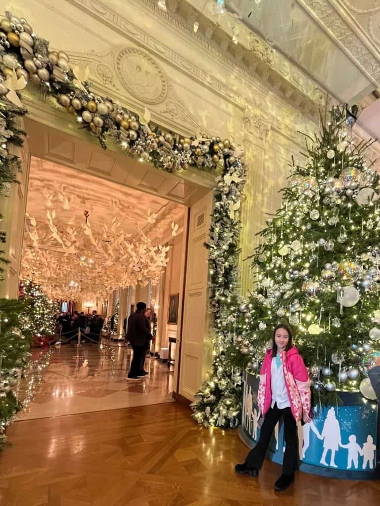 A smiling girl in a pink jacket stands in front of a 12 foot decorated Christmas tree and a hallway with a ceiling decorated with doves on a White House tour.