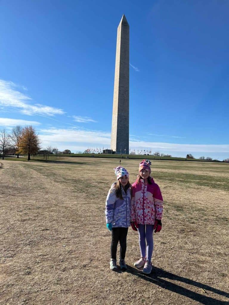 Two young girls bundled in winter coats and hats standing in front of the Washington Monument.