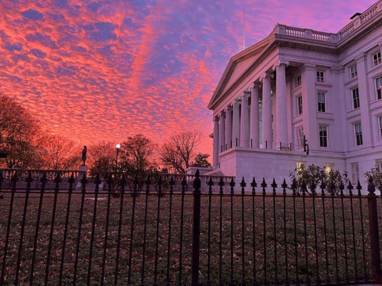 White government building with an iron gate and green lawn at dusk in Washington DC with a colorful pink and blue sky.
