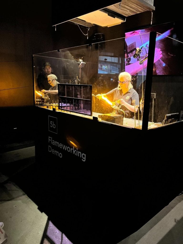 Glassblowing artist shaping molten glass behind safety glass during a live demo at the Corning Museum of Glass.