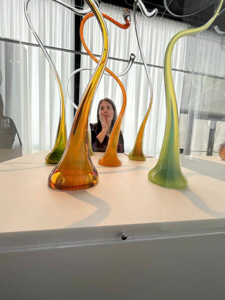 Woman stands next to whimsical glass sculptures in bright green and orange at the Corning Museum of Glass.