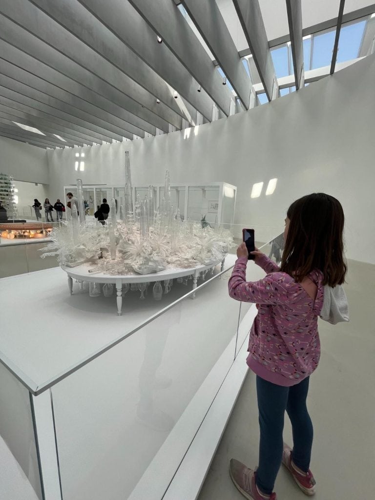 Is Corning Museum of Glass good for kids? A young girl photographs an elaborate white glass sculpture in a sunlit gallery, proving the answer is yes.