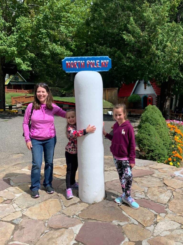 Our first tiny adventure in the Adirondacks as a family. A mom and two girls smiling next to North Pole, NY icy post tourist attraction in New York.
