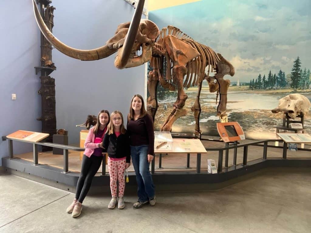 Visiting an interactive museum on a family day trip. A woman reclaiming family time on a summer day off with her two girls, standing in front of a Wooly Mammoth skeleton in an Ithaca, NY museum.