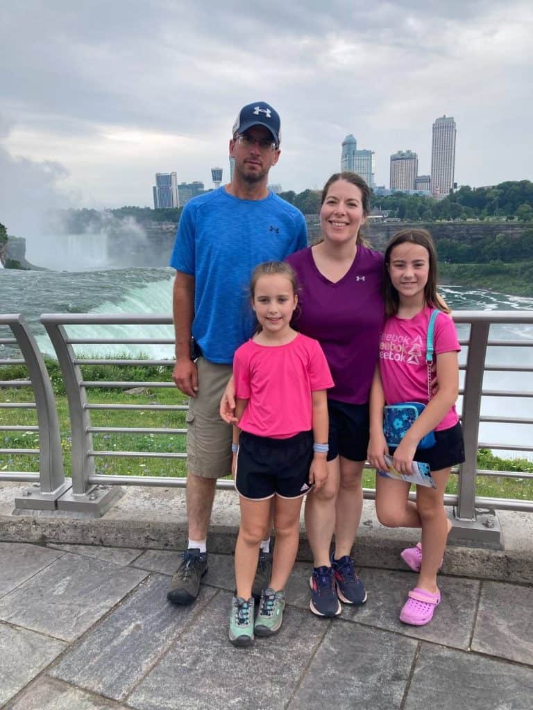 Our family of 4 taking an iconic bucket list trip in NY to see Niagara Falls. A family of 4 in shorts on a bridge overlooking Niagara Falls, NY waterfalls.
