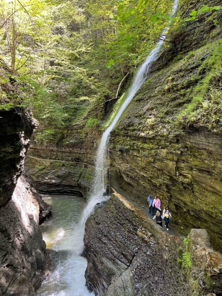 Showing the path that families with kids will walk to go underneath waterfalls at Watkins Glen, NY. A large waterfall cascading down rock formation, with people walking on a path below it and under it.