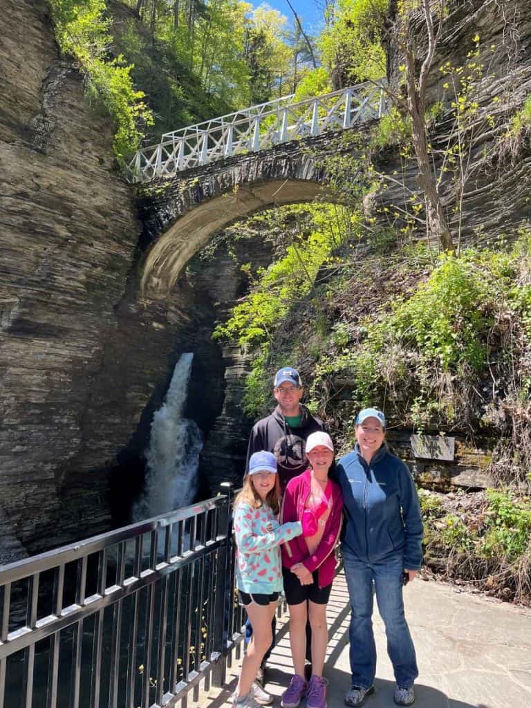 A family of 4 takes a photo in front of a waterfall on a hike at Watkins Glen, NY state park. A family of 4 stands in front of a waterfall on a stone entrance pathway at Watkins Glen state park. A bridge is overhead.