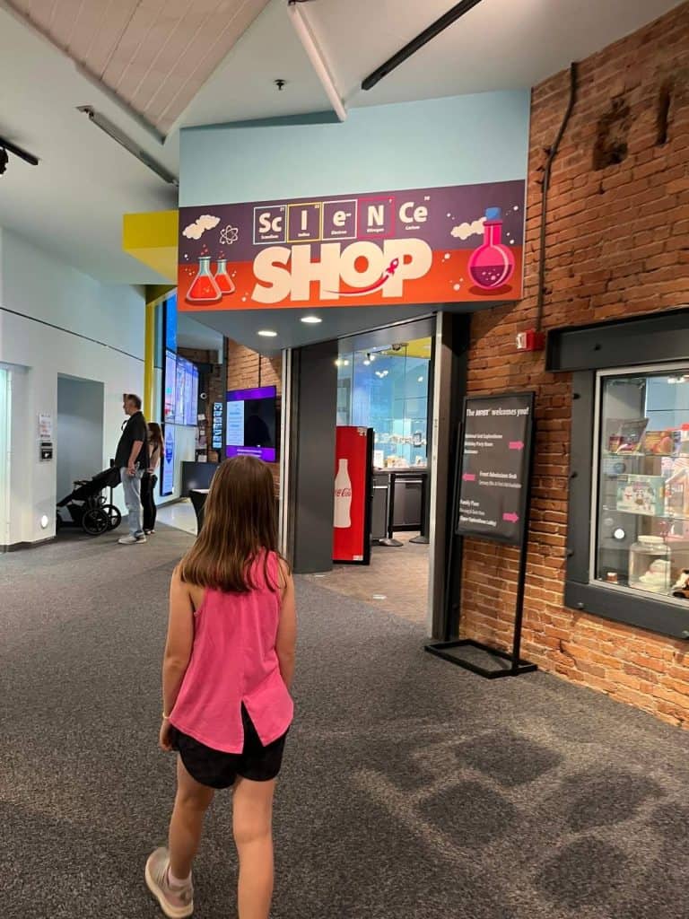 A girl walks into the gift shop at the MOST science museum in Syracuse, NY. A girl walks toward an alcove with brick exterior that has a Science Shop sign with chemical bottle graphics.