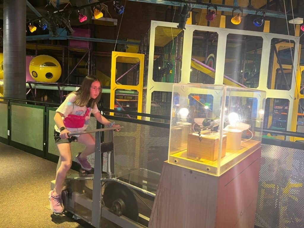 A girl rides an indoor bike at a science exhibit and learns how that energy can be used to light up a lightbulb. A girl rides an indoor bike that lights up a lightbulb in a clear case in front of the bike.
