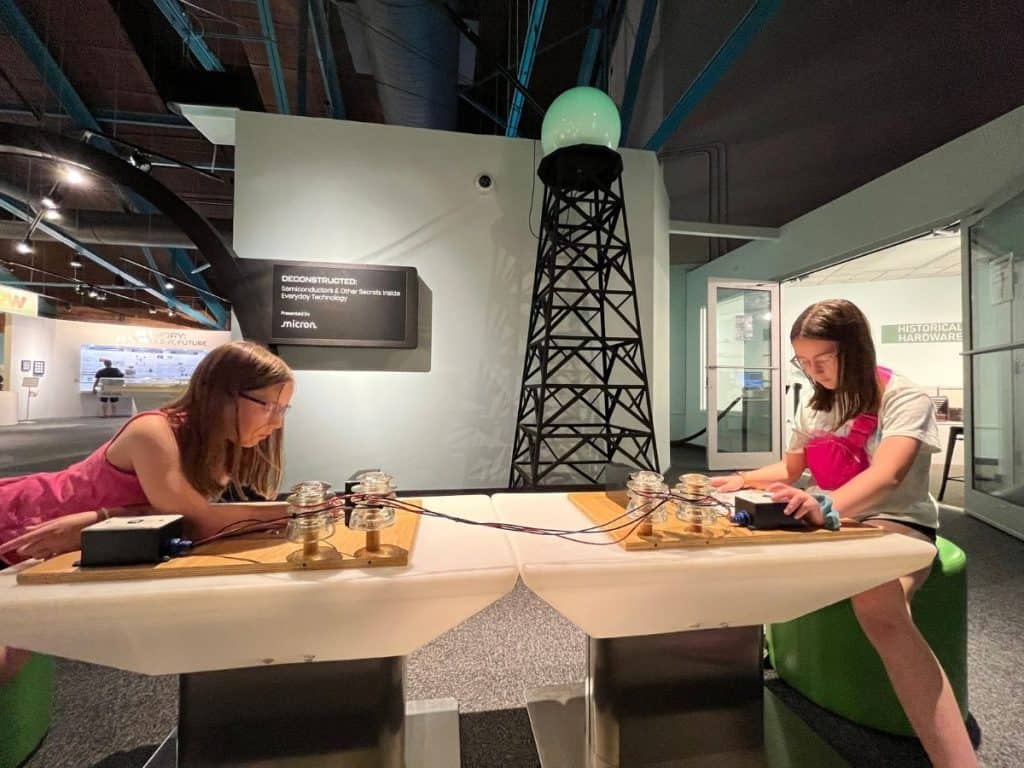 Two girls practice communicating with Morse Code in a science exhibit at the MOST museum in Syracuse, NY. Two girls sit on either end of a table at a Morse Code station in front of a replica communications tower.