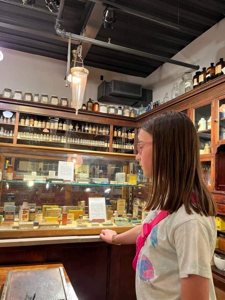 A girl looks at potions, lotions, and tonics in the apothecary replica of the Syracuse science museum. A girl looks at a clear case with bottles and jars showing medicines and lotions, with wooden displays along the walls of glass jars with tonics and potions.